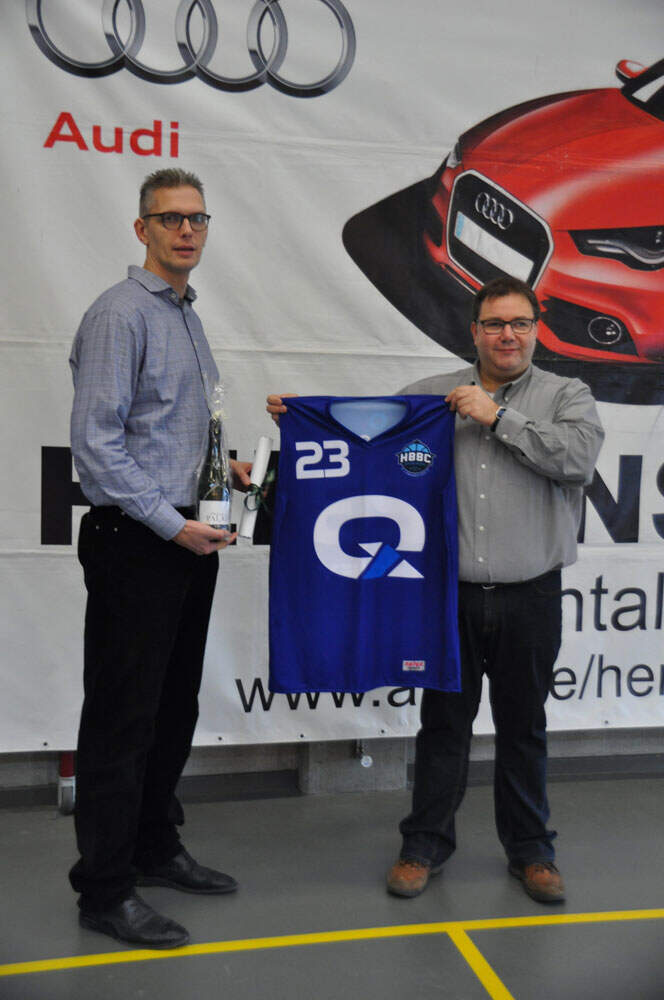 QCIFY blue uniform being held between CTO, Bert Peelaers and another man both wearing grey collared shirts and black pants wearing glasses in front of Audi sponsorship banner